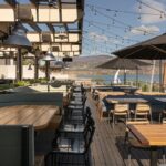 A stylish outdoor dining area with wooden tables, chairs, and umbrellas, overlooking a lake and mountains, illuminated by string lights.