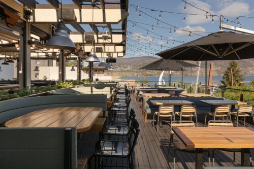 A stylish outdoor dining area with wooden tables, chairs, and umbrellas, overlooking a lake and mountains, illuminated by string lights.