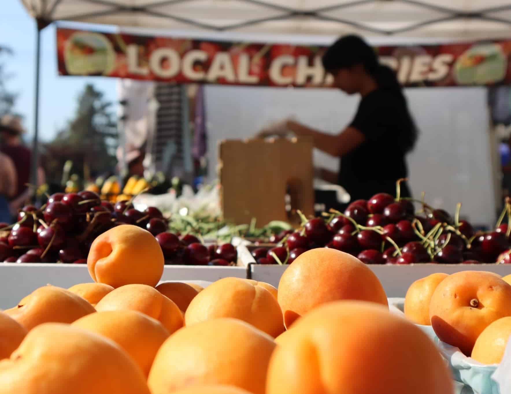 A variety of fresh peaches and cherries are displayed at a market stall, with a person blurred in the background. A banner reading "LOCAL CHERRIES" is visible above.