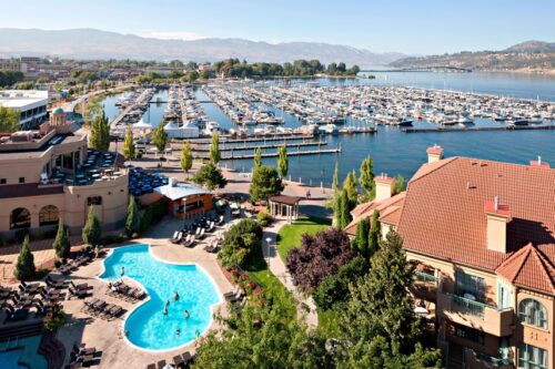 A scenic view featuring a pool area and a marina filled with boats, surrounded by greenery and buildings under a clear blue sky.