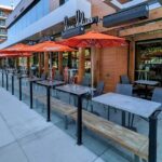 A restaurant patio featuring several tables with orange umbrellas, surrounded by a glass railing and wooden posts, under a modern building.