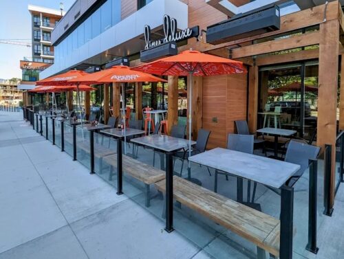 A restaurant patio featuring several tables with orange umbrellas, surrounded by a glass railing and wooden posts, under a modern building.