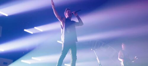 A musician performing on stage with a microphone, surrounded by dramatic lighting and smoke effects.