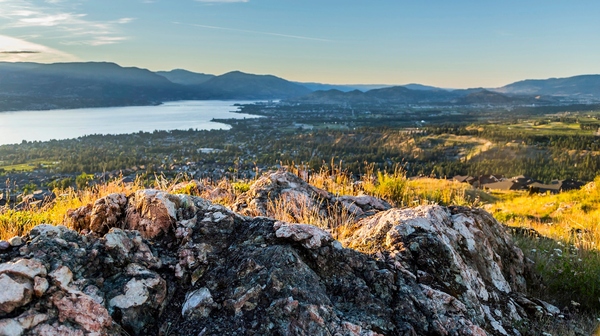 A scenic view of a valley and lake surrounded by mountains, with rocky foreground and grassy patches.