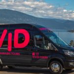 A black tour van with bright pink lettering displaying "VIVID" and the words "WINE • BEER • CANNABIS TOURS," parked near a scenic view of a lake and mountains.