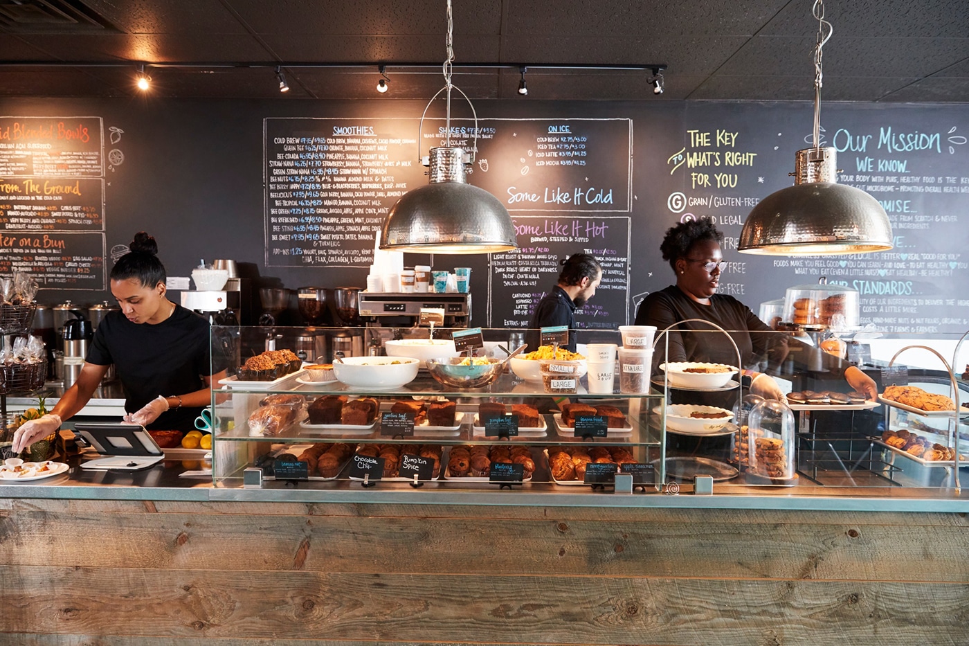 A café interior featuring a counter with baked goods, a menu board, and two employees preparing food and drinks.