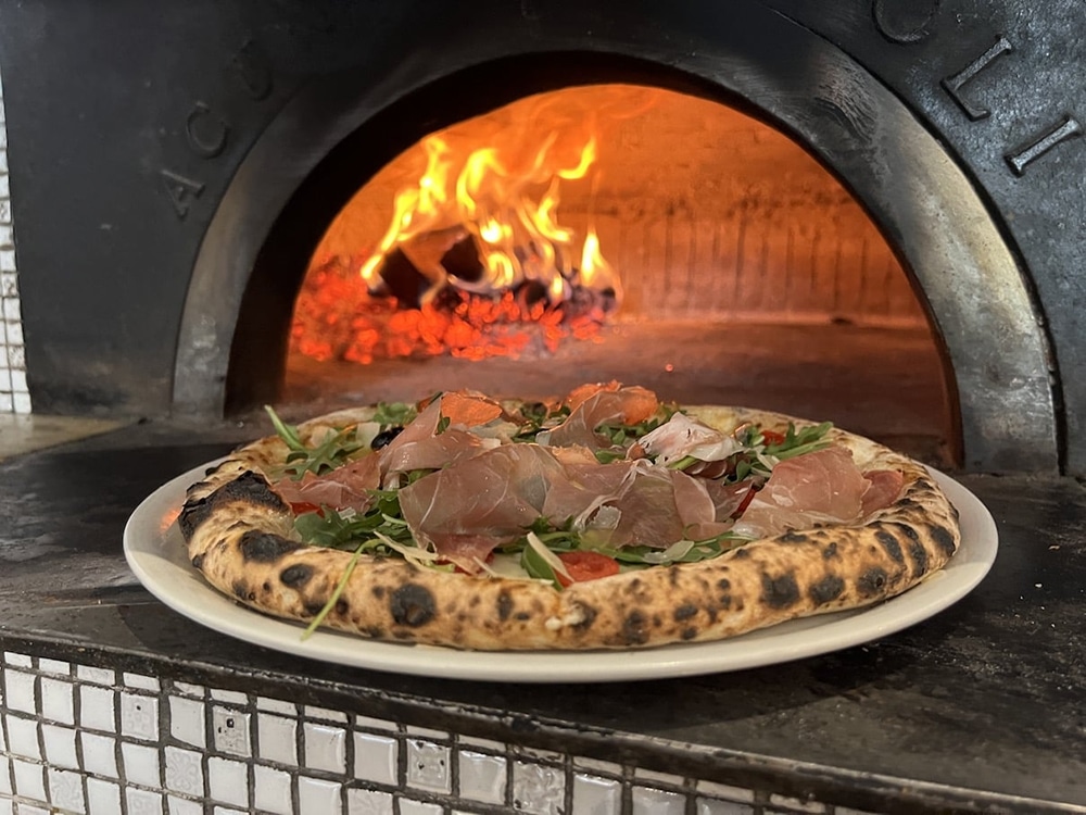 A freshly baked pizza topped with greens and slices of prosciutto, placed in front of a wood-fired oven with flames visible.