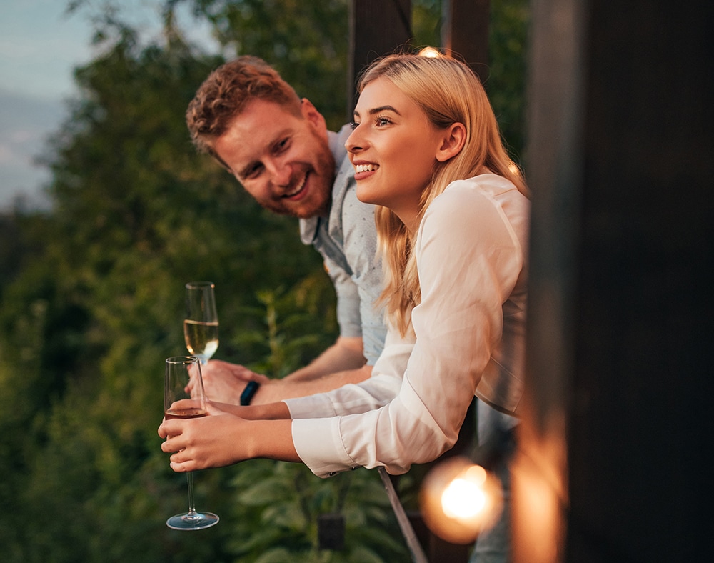 A man and a woman are smiling while holding champagne glasses on a balcony, surrounded by greenery.