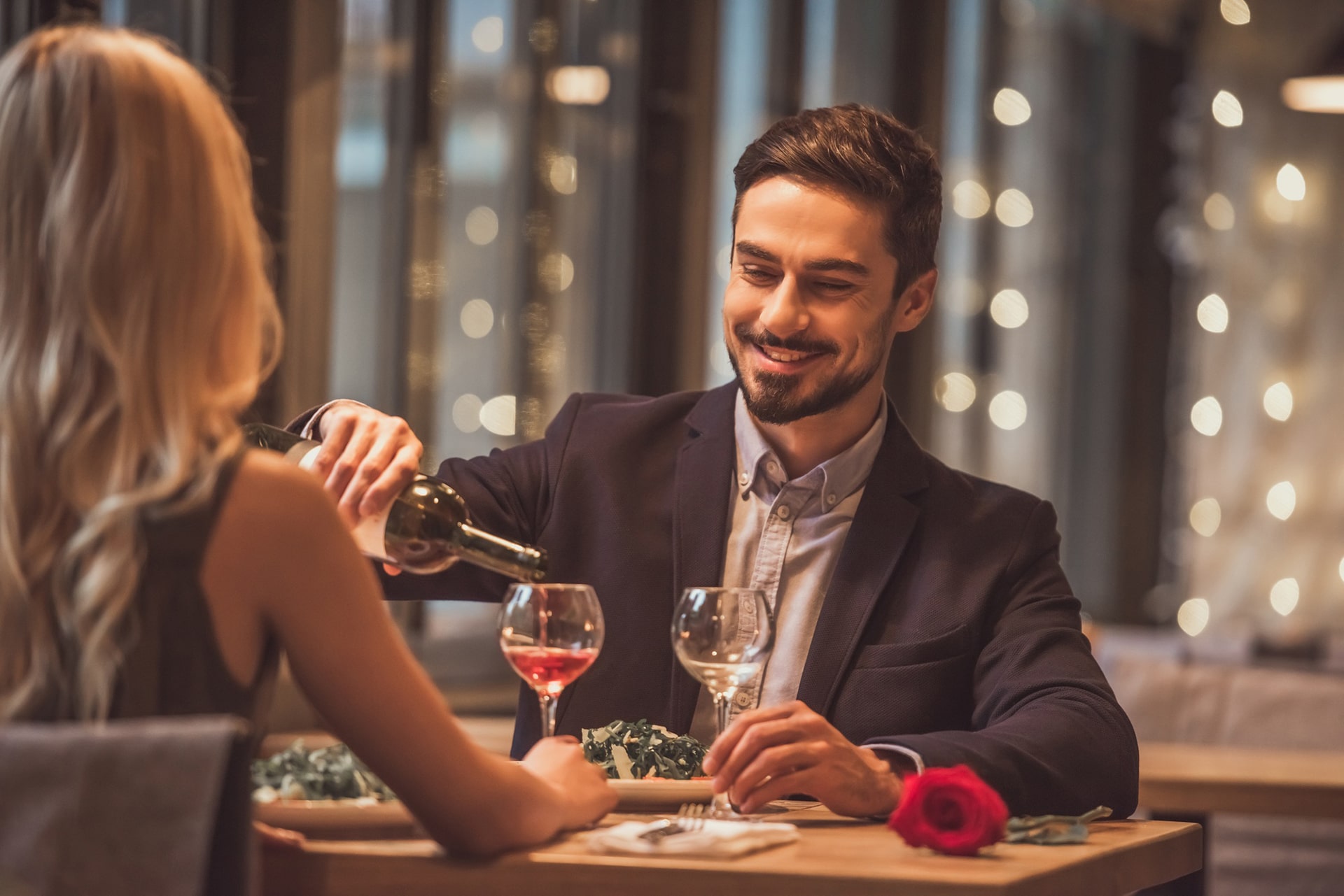 A man in a suit is pouring wine into a glass for a woman at a table, which has a romantic atmosphere with soft lighting and a red rose.