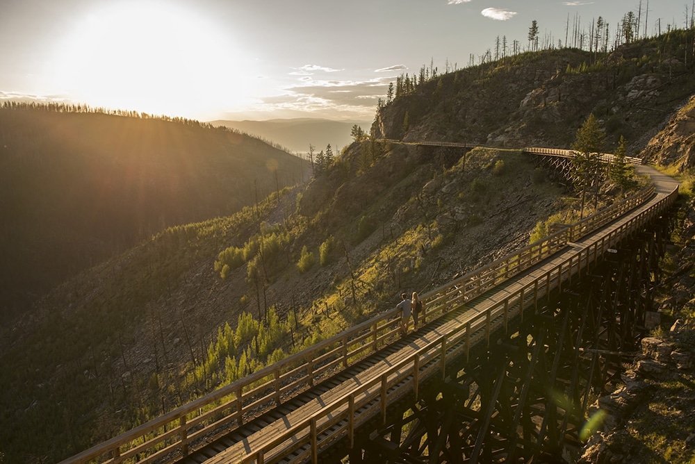A scenic view of a wooden walking path winding along a hillside at sunset, with two people walking on it and trees in the background.