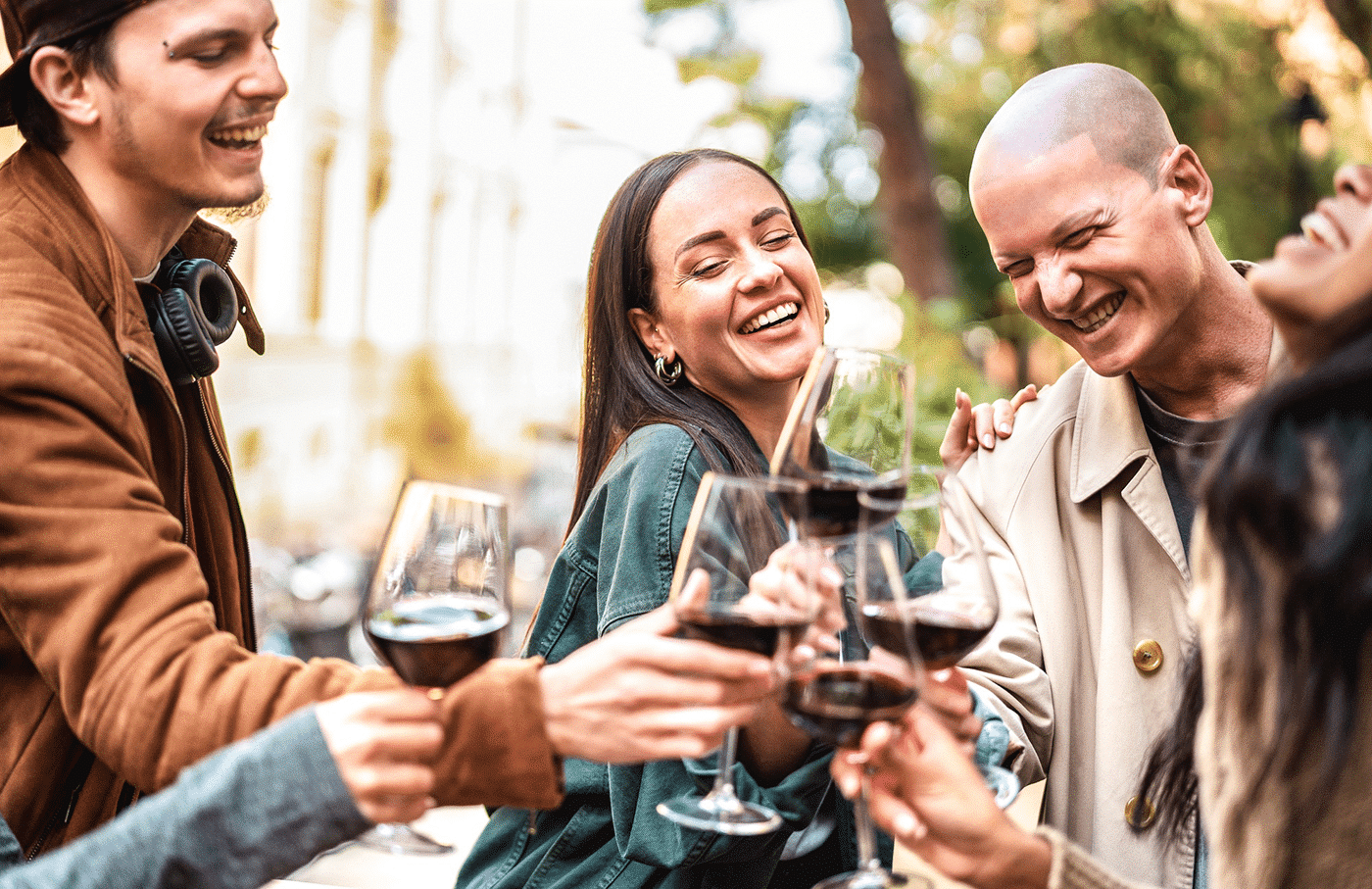 A group of five friends smiling and toasting with glasses of red wine outdoors.