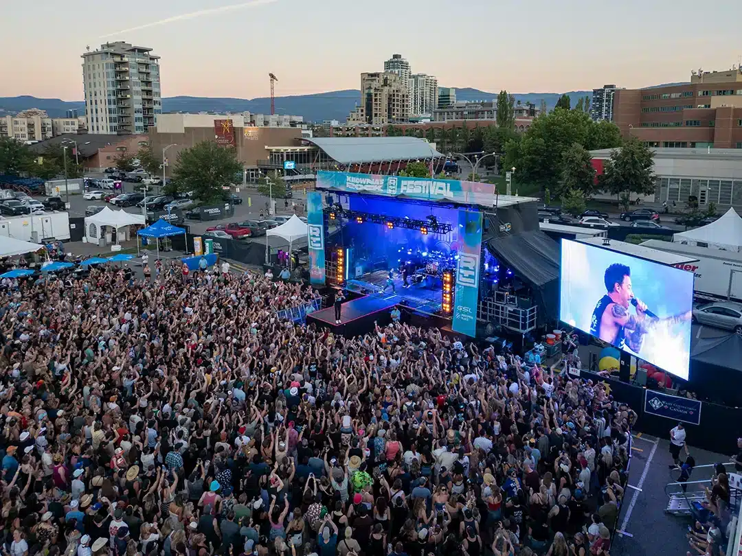 A large crowd attends a music festival, with a performer on stage and a big screen showing a close-up of the artist, set against a backdrop of buildings and trees.