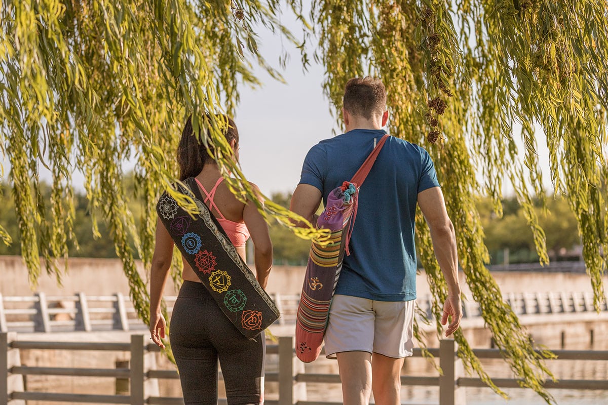 A woman and a man walking near a body of water, carrying yoga mats, under a willow tree.